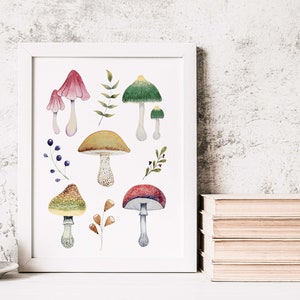 May include: A framed watercolor print featuring various colorful mushrooms and foliage. The artwork includes mushrooms with red, green, and brown caps, along with sprigs of leaves and berries. The print is displayed on a white surface.