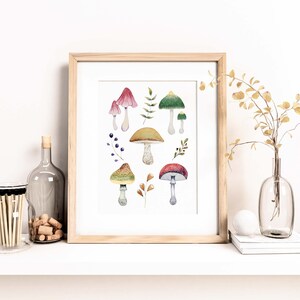 May include: A framed watercolor print featuring various colorful mushrooms and foliage. The mushrooms have red, green, yellow, and brown caps with white stems. The artwork is displayed on a white shelf with decorative items.