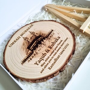 May include: A wooden round plaque with a laser-engraved image of the Kaaba, a mosque in Mecca, Saudi Arabia. The plaque also includes the text "Umrah Mubarak Yaqub & Rubaka May Allah accept your Umrah and allow you to visit many more times Ramadan 1444/2023".