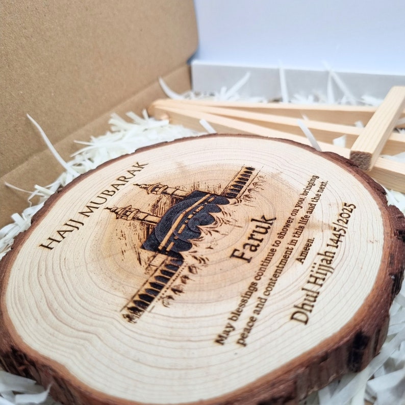 May include: A wooden round plaque with a laser-engraved image of the Kaaba, a mosque, and the text "Hajj Mubarak Faruk May blessings continue to shower on you, bringing peace and contentment in this life and the next. Ameen. Dhul Hijjah 1445/2025".