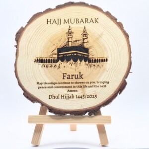 May include: A wooden round plaque with a laser-engraved image of the Kaaba, a mosque, and the text "HAJJ MUBARAK". The plaque also includes the name "Faruk" and the text "May blessings continue to shower on you, bringing peace and contentment in this life and the next. Ameen. Dhul Hijjah 1445/2025".