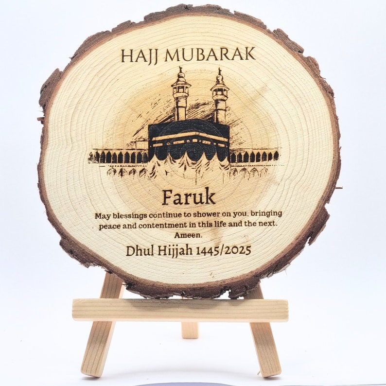 May include: A wooden round plaque with a laser-engraved image of the Kaaba, a mosque, and the text "HAJJ MUBARAK". The plaque also includes the name "Faruk" and the text "May blessings continue to shower on you, bringing peace and contentment in this life and the next. Ameen. Dhul Hijjah 1445/2025".