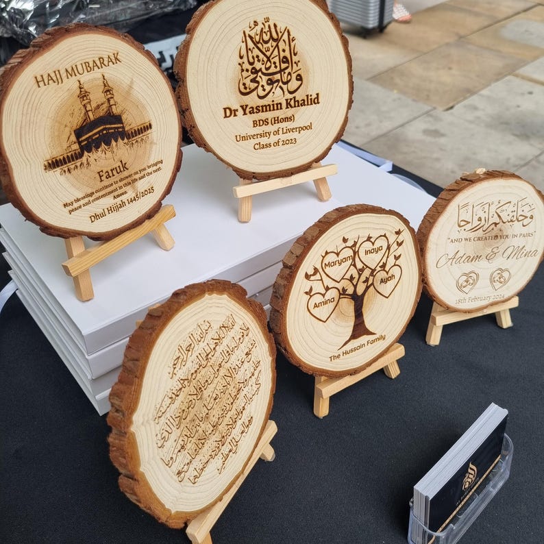 May include: Six wooden plaques with engraved Arabic calligraphy and personalized messages. The plaques are mounted on small wooden stands. The messages include a Hajj Mubarak greeting, a graduation message for Dr. Yasmin Khalid, a family tree with names, and a wedding message for Adam & Mina.