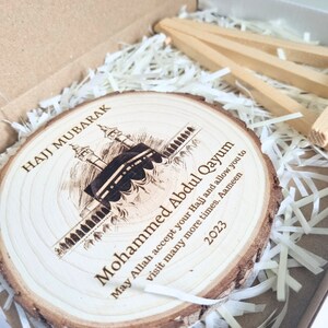 May include: A wooden round plaque with a laser-engraved image of the Kaaba in Mecca, Saudi Arabia. The plaque has the text "Hajj Mubarak" and "Mohammed Abdul Qayum" with a message wishing for a successful Hajj pilgrimage and many more visits to the holy site. The year 2023 is also engraved on the plaque.