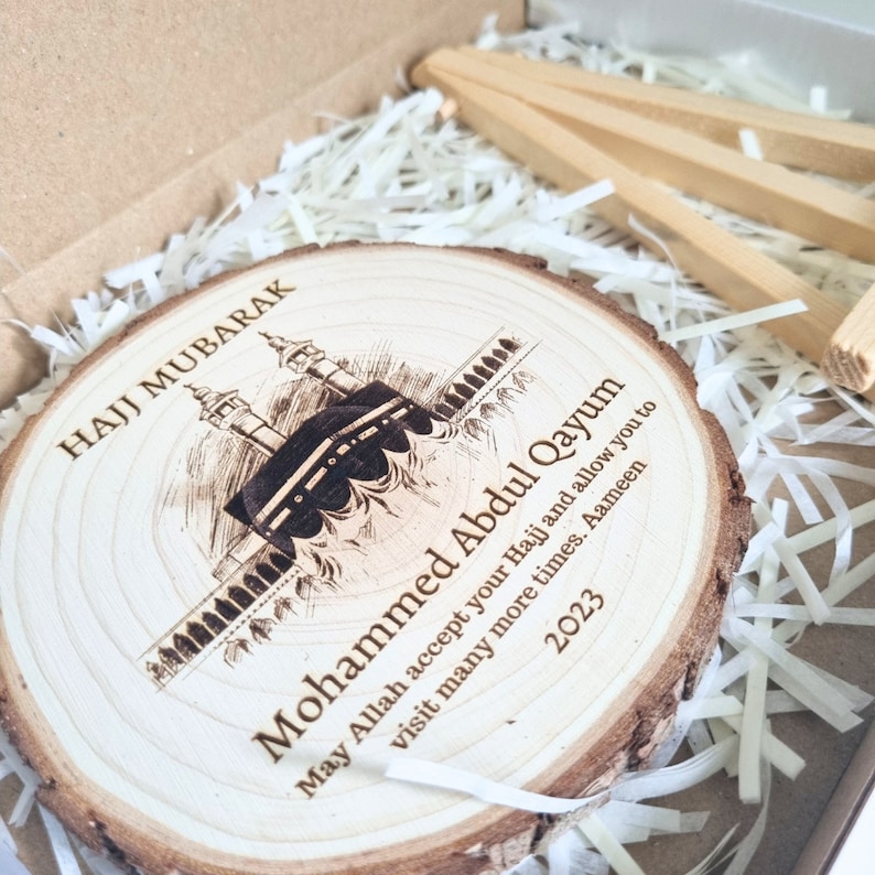 May include: A wooden round plaque with a laser-engraved image of the Kaaba in Mecca, Saudi Arabia. The plaque has the text "Hajj Mubarak" and "Mohammed Abdul Qayum" with a message wishing for a successful Hajj pilgrimage and many more visits to the holy site. The year 2023 is also engraved on the plaque.