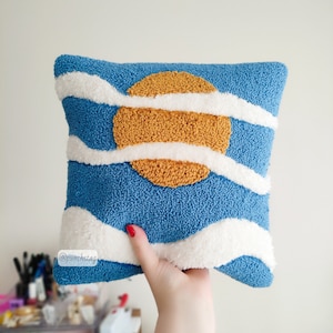 Könnte beinhalten: Ein blaues und weißes handgenähtes Kissen mit einem gelben Sonnendesign. Das Kissen wird von einer Hand hochgehalten. Der Text "@punchstag" ist auf dem Kissen sichtbar.