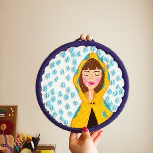 May include: A needlepoint embroidery hoop with a colorful image of a person wearing a yellow raincoat and standing in the rain. The background is white with blue raindrops.