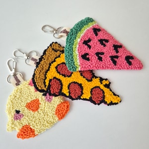May include: Three food-shaped keyrings. One is a watermelon slice with a green rind, pink flesh, and black seeds. Another is a pizza slice with yellow cheese and red pepperoni. The last is a yellow and orange cake slice.
