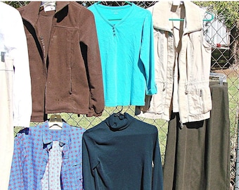 May include: Assortment of clothing items hanging on a fence.  The items include a white long-sleeved shirt, a brown jacket, a turquoise long-sleeved shirt, a beige vest, a blue and red patterned shirt, a dark green turtleneck sweater, and a brown skirt.