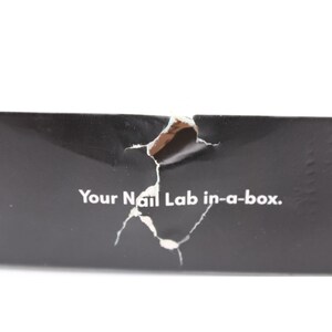 Op de afbeelding: Close-up van een zwarte doos met een gescheurd gedeelte dat een lichtere kleur eronder onthult. De tekst "Your Nail Lab in-a-box." is in het wit op de doos gedrukt.