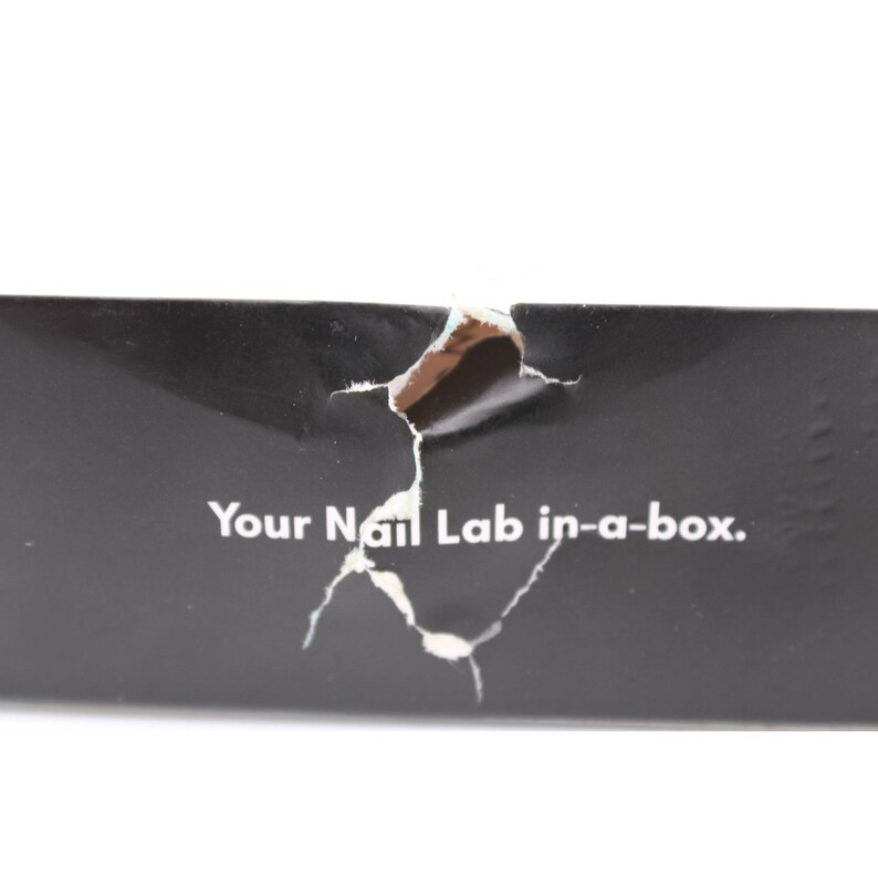 Op de afbeelding: Close-up van een zwarte doos met een gescheurd gedeelte dat een lichtere kleur eronder onthult. De tekst "Your Nail Lab in-a-box." is in het wit op de doos gedrukt.