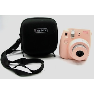 May include: A pink Fujifilm Instax Mini 9 instant camera with a black carrying case and a strap. The camera has a white lens ring and a black lens cover. The case has the word "instax" printed on it.