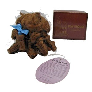 May include: A brown wig with curly hair and a blue ribbon. The wig is packaged in a brown box with the text "PLAYHOUSE Collection" on it. The wig is also labeled with the text "PLAYHOUSE Collection" and "MADE IN KOREA".