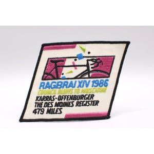 May include: A vintage embroidered patch with a bicycle graphic in pink and black. The patch features the text "RAGBRAI XIV 1986" and "479 MILES" in blue and white. The patch has a black border and a cream background.