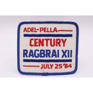 May include: A square patch with a white background and a blue border. The patch features the text "ADEL-PELLA", "CENTURY", "RAGBRAI XII", and "JULY 25 '84" in red and blue lettering. The patch is likely a commemorative item.