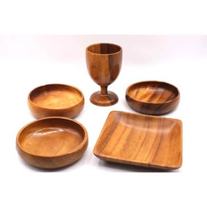 Puede incluir: Un conjunto de piezas de servicio de madera, que incluye cuencos y una copa. Los artículos están hechos de madera marrón claro con patrones de vetas visibles. El conjunto incluye cuencos redondos y un plato cuadrado.