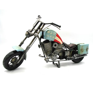 May include: A metal motorcycle sculpture with a light blue and red American flag design on the gas tank. The sculpture has a black tire, a light blue front fender, and a green storage box.