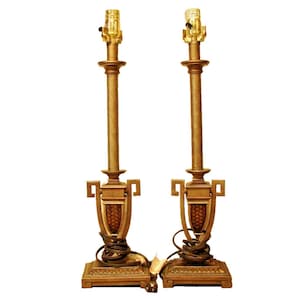 May include: A pair of gold table lamps with a classic design. Each lamp features a tall, slender base with a decorative urn-shaped top and a simple, clear glass shade.