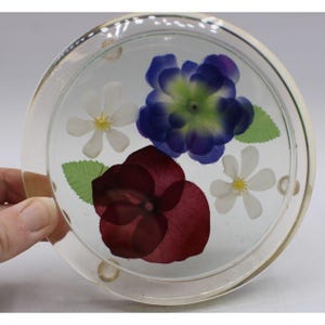 May include: A circular clear resin coaster featuring embedded pressed flowers including a large deep purple bloom, a smaller blue flower with a green center, and two white blossoms with yellow centers, accented by two small green leaves.