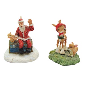 May include: Two Christmas ornaments, one depicting Santa Claus sitting on a blue and white chest with a small white kitten, and the other depicting a red-hooded deer with a smaller deer beside it. Both ornaments are on a white base.