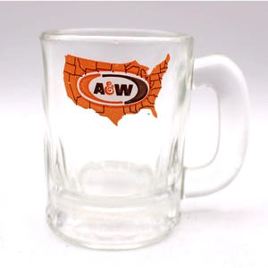 May include: Clear glass mug with a handle, featuring an orange and brown graphic of the United States with the A&W logo. The mug has a rectangular shape and is likely for beverages.