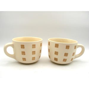 May include: Two cream-colored ceramic mugs with rounded handles. Each mug is decorated with a pattern of evenly spaced, light brown squares. The mugs are set against a plain white background, highlighting their simple design.
