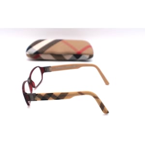 May include: Eyeglasses with a burgundy frame and tan arms featuring a black plaid pattern. A matching plaid eyeglasses case is in the background. The glasses have a rectangular shape.
