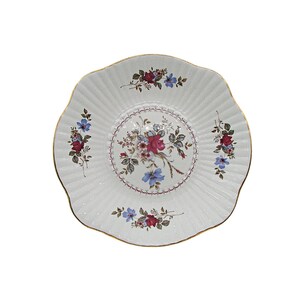 May include: White porcelain plate with a scalloped edge and gold trim. The plate features a central floral design with red and blue flowers, surrounded by a decorative border. Additional floral clusters are placed around the plate's rim.
