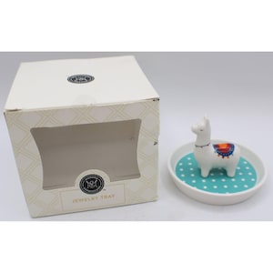 May include: A white ceramic jewelry tray featuring a small, standing white llama figurine with a colorful saddle. The tray has a teal base with white polka dots. The item is displayed next to its original white box labeled Modern Expressions.