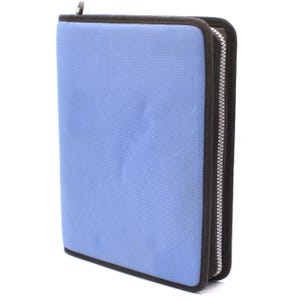 May include: A light blue zippered binder with a black trim and a silver zipper. The binder is rectangular and appears to be made of a textured fabric. A silver metal loop is at the top.