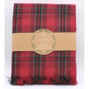 May include: An oblong tablecloth featuring a red, green, and black plaid pattern with a fringed edge. A tan paper band wraps around the center, printed with the text ASPEN EST. 1949 OBLONG TABLECLOTH 60 x 102 (152CM x 259CM) and a leaf icon.