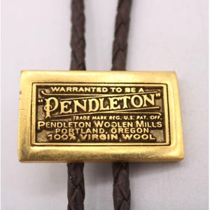May include: A bolo tie featuring a rectangular gold-toned metal slide with the Pendleton logo and text. The slide is attached to a dark brown braided leather cord. The text on the slide reads "WARRANTED TO BE A PENDLETON".