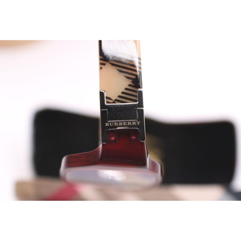 May include: Close-up of a pair of Burberry sunglasses. The frame is a deep red color, with a silver hinge bearing the Burberry logo. The arm features a beige and black plaid pattern.