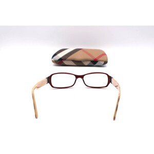 May include: Eyeglasses with a burgundy frame and light-colored arms. The glasses are positioned in front of a matching case with a plaid pattern in tan, red, and black. The glasses have a rectangular shape.
