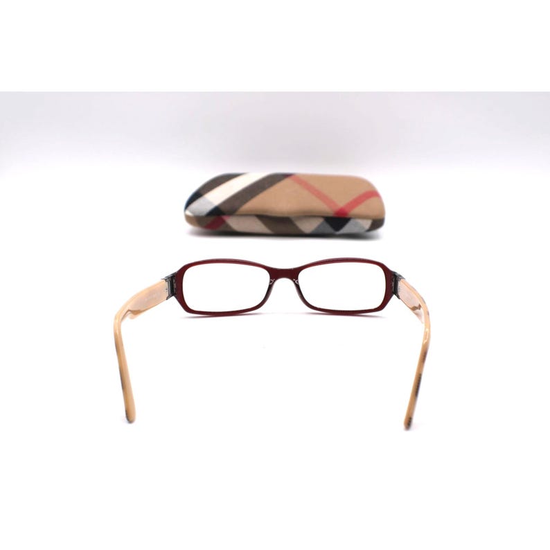May include: Eyeglasses with a burgundy frame and light-colored arms. The glasses are positioned in front of a matching case with a plaid pattern in tan, red, and black. The glasses have a rectangular shape.