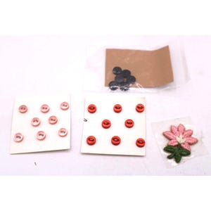 May include: A collection of sewing notions including pink and red buttons, and a pink flower patch with green leaves. A small bag of black buttons is also included.