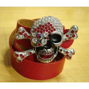 May include: A red leather belt with a silver skull and crossbones buckle. The skull is decorated with red and clear rhinestones.