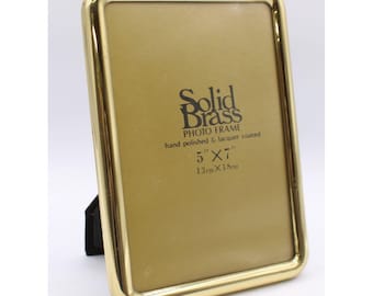 May include: A rectangular solid brass photo frame with a polished gold finish. The frame features rounded corners and a black easel back. Printed on the center insert is the text Solid Brass PHOTO FRAME hand polished & lacquer coated 5" x 7".