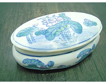 May include: An oval ceramic trinket box with a lid. The box is decorated with a blue and white floral design featuring lotus flowers and leaves. The box is white with a blue and green floral pattern.