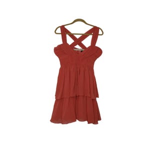 May include: Coral-colored dress with a tiered skirt and crisscross straps. The dress is hanging on a wooden hanger. The fabric appears to be lightweight and flowing, suitable for warm weather.