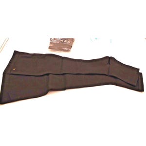 May include: A folded black garment, possibly a sleeve or leg warmer, lies on a white surface. A small, sealed package is visible in the background. A small gold-colored emblem is on the garment.