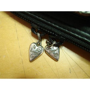 Puede incluir: Primer plano de una cremallera negra con dos colgantes en forma de coraz&oacute;n. Uno dice "Love" con un dise&ntilde;o floral, y el otro dice "Amour". Los colgantes son plateados y cuelgan de la cremallera.