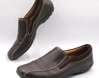 May include: Two brown leather slip-on shoes with white stitching. The shoes have a rounded toe and a dark brown sole. One shoe is positioned in the background, and the other is in the foreground. The shoes appear to be new.