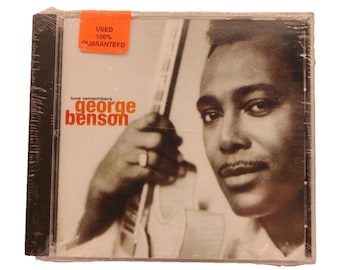 May include: A CD case featuring George Benson, with the album title "love remembers" and the artist's name in orange. The image is in black and white, with a sticker that says "USED 100% GUARANTEED". The CD is sealed in plastic.