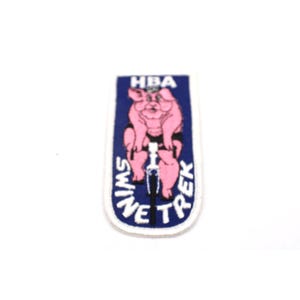 May include: A rectangular embroidered patch with a dark blue background and a white border. The patch features a pink pig riding a bicycle, with the text HBA at the top and SWINE TREK written in white capital letters along the bottom curve.