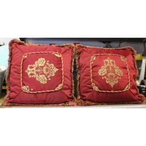 May include: Two square red throw pillows featuring a central panel with a gold floral damask pattern. Each pillow is framed by a decorative twisted rope trim and finished with a matching red fringe along the outer edges of the square cushions.