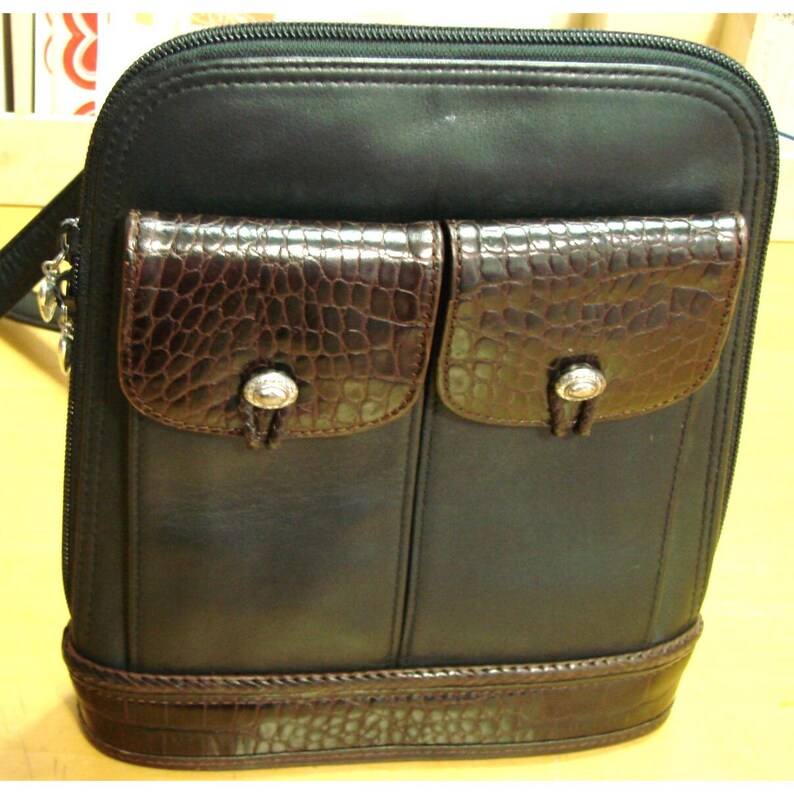 Puede incluir: Bolso bandolera de cuero negro con dos bolsillos delanteros con dise&ntilde;o marr&oacute;n con textura de cocodrilo. El bolso tiene cierre de cremallera y base marr&oacute;n con textura de cocodrilo. Detalles plateados en los bolsillos.