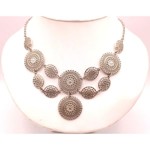 May include: Silver-toned necklace with a series of circular and oval decorative elements. The necklace is displayed on a white mannequin bust. The design features intricate patterns and textures, creating a detailed and elegant appearance.