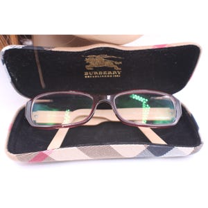 May include: Open eyeglasses case with Burberry logo and glasses. The glasses have a dark red frame and clear lenses. The case is black inside with a tan and plaid exterior.