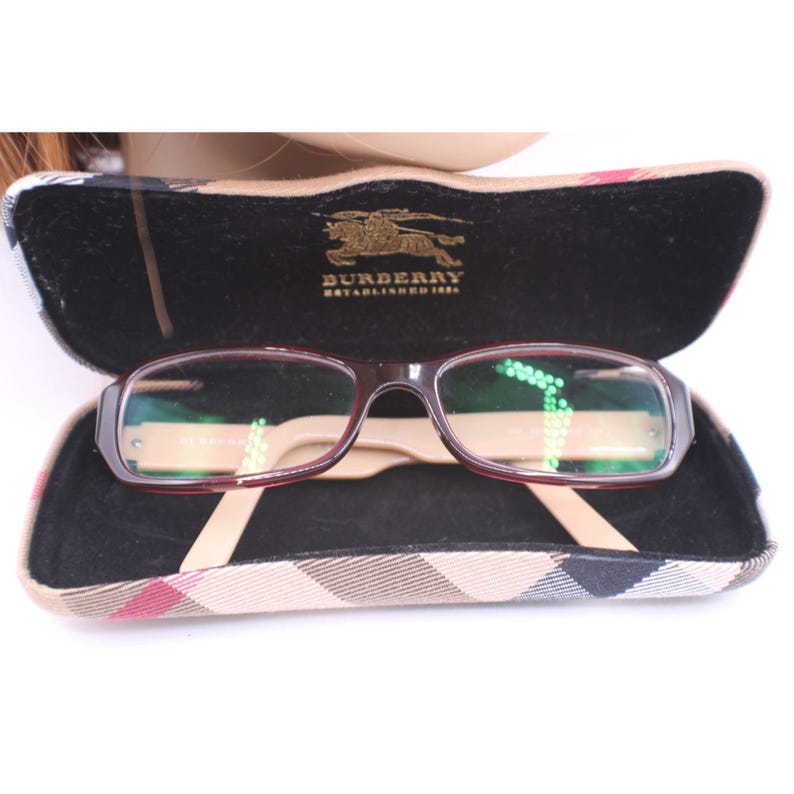May include: Open eyeglasses case with Burberry logo and glasses. The glasses have a dark red frame and clear lenses. The case is black inside with a tan and plaid exterior.